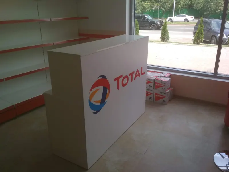 Белая стойка с логотипом TOTAL у окна, у стены стеллажи