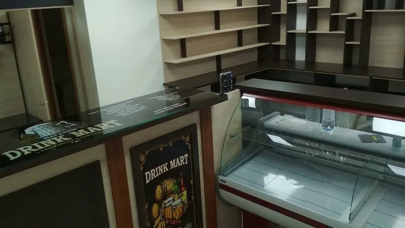 Стойка со стеклянной столешницей и постером DRINK MART в раме, рядом холодильная витрина, за ней стеллажная стена