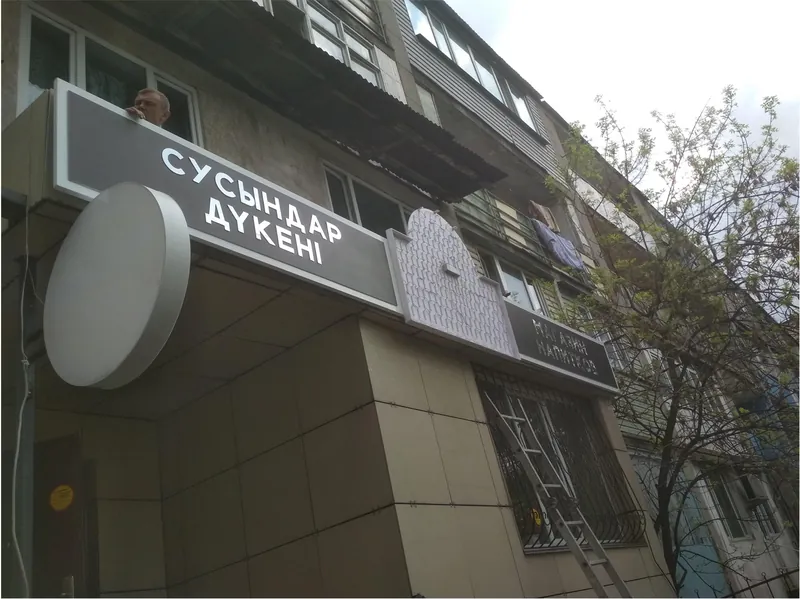 Монтаж вывески на фасаде магазина: надпись «Сусындар дүкені» и круглый короб без логотипа
